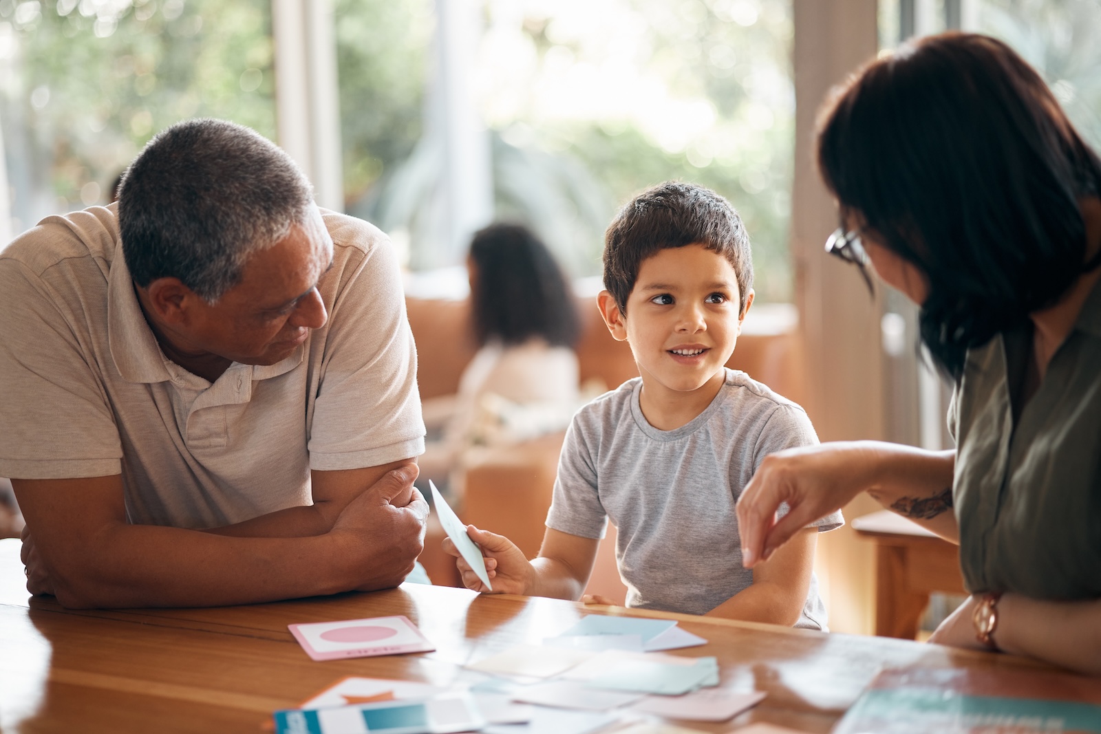 A young boy smiling during a family meeting at a wooden table with his father and mother in a bright, sunlit room.