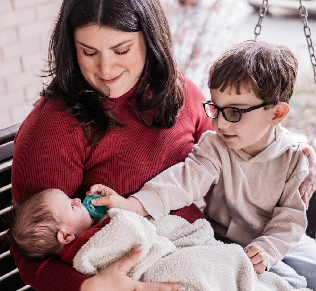 Deena from DFN Connected Growth Coaching sits on a porch swing with her infant and older son, sharing a tender family moment.