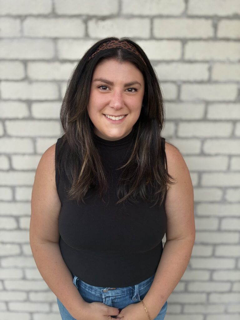 Full-length professional headshot of Deena, lead coach at DFN Connected Growth Coaching, wearing a black sleeveless top and jeans against a white brick wall.